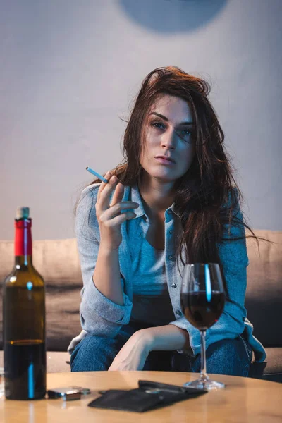 Drunk woman holding cigarette while sitting near red wine and empty wallet on blurred foreground — Stock Photo