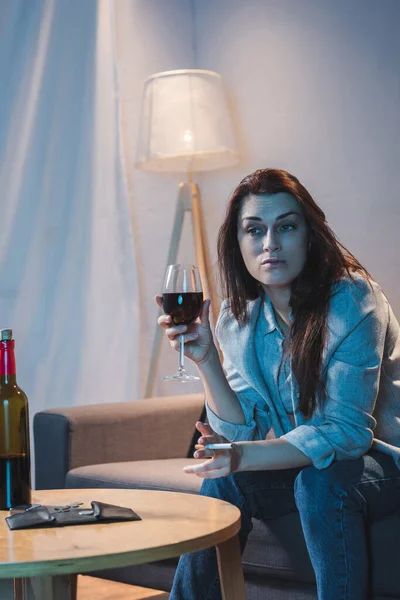 Frustrated woman holding cigarette and glass of red wine near bottle and empty wallet on table — Stock Photo