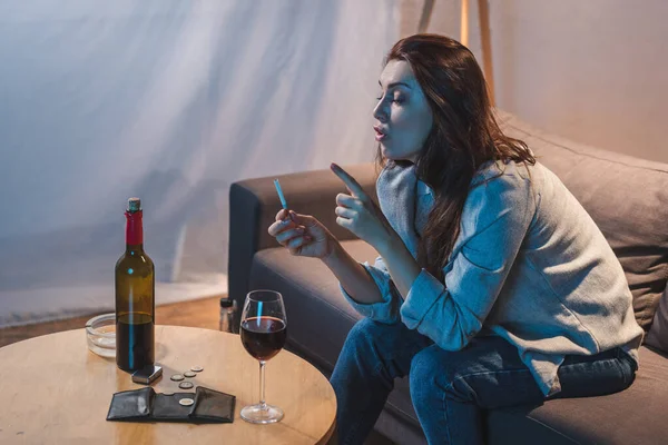 Mulher bêbada segurando cigarro enquanto sentado perto de vinho tinto, moedas e carteira vazia na mesa — Fotografia de Stock