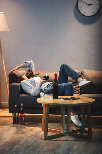 Betrunkene Frau liegt auf Sofa mit Weinglas neben Flaschen auf Tisch und Boden — Stockfoto