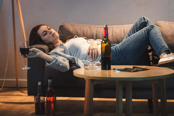 Mujer borracha acostada en el sofá con copa de vino cerca de botellas en el suelo y la mesa - foto de stock