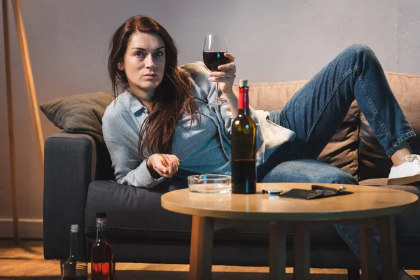 Drunk woman with glass of red wine lying on sofa near bottles of alcohol — Stock Photo