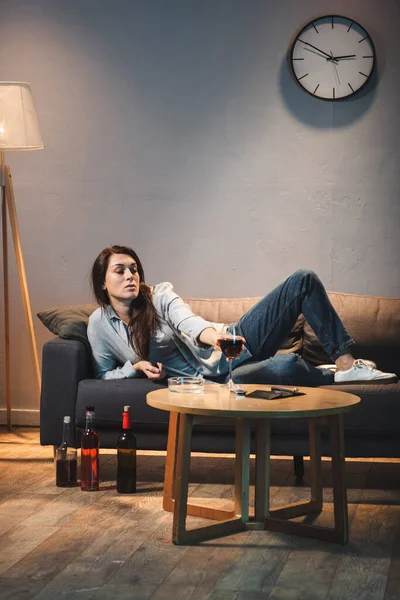Lonely woman lying on sofa near bottles of alcohol on floor and glass on table — Stock Photo