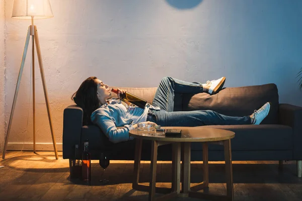 Seitenansicht einer depressiven Frau, die Wein aus einer Flasche trinkt, während sie auf dem Sofa liegt — Stockfoto