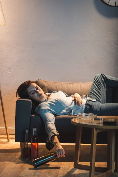 Drunk woman sleeping on sofa with glass of red wine near bottles on floor — Stock Photo