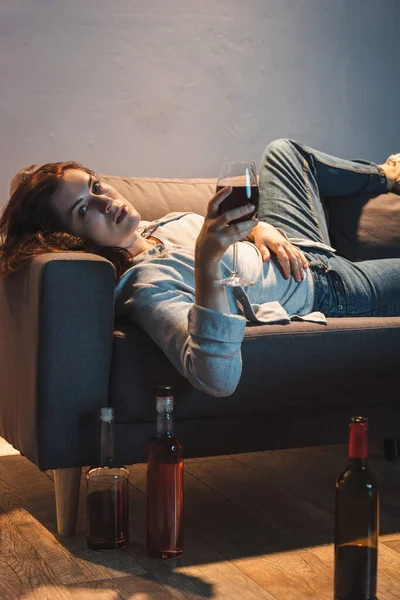 Lonely woman looking at camera while lying on sofa with glass of red wine near bottles on floor — Stock Photo