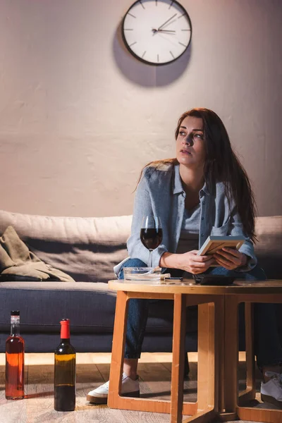 Mujer deprimida mirando hacia otro lado mientras está sentada cerca de botellas de alcohol y sosteniendo el marco de fotos - foto de stock