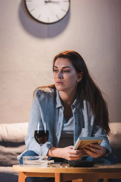 Upset, alcohol-addicted woman holding photo frame while sitting near glass of red wine — Stock Photo