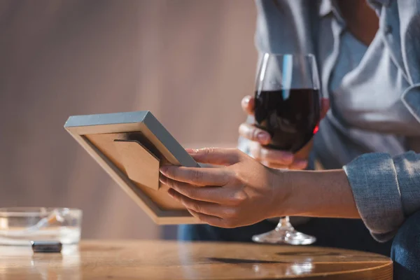 Cropped view of woman holding glass of red wine and photo frame on blurred background — Stock Photo