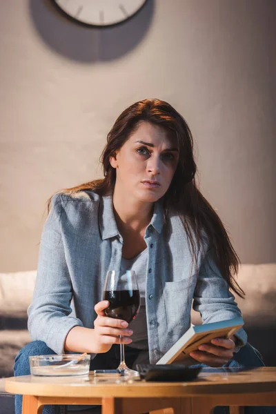 Mujer deprimida mirando a la cámara mientras está sentada con un vaso de vino tinto y sosteniendo el marco de la foto - foto de stock