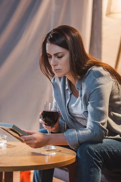 Mujer molesta y frustrada mirando el marco de la foto y sosteniendo una copa de vino tinto - foto de stock