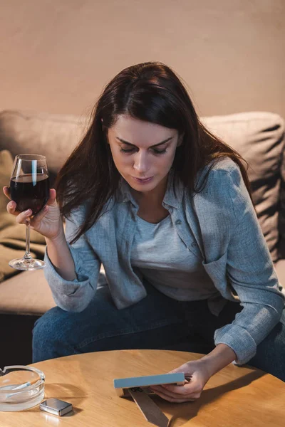 Femme triste regardant cadre photo tout en étant assis à la maison seul avec un verre de vin rouge — Photo de stock