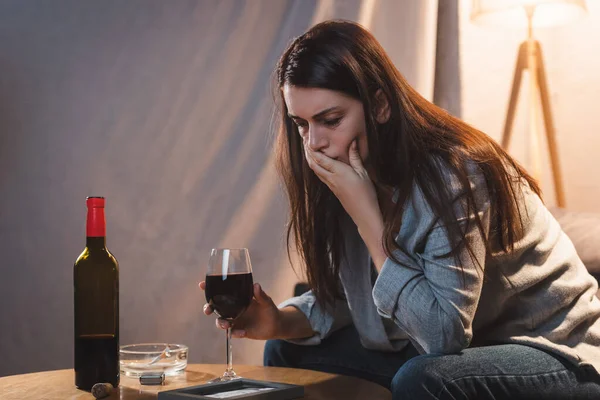 Mulher deprimida com copo de vinho cobrindo a boca com a mão enquanto olha para moldura da foto — Fotografia de Stock