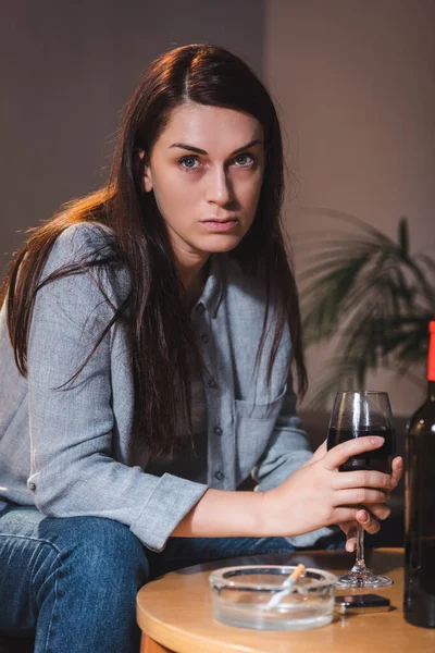 Mujer alcohólica mirando hacia otro lado mientras está sentada en casa sola con una copa de vino tinto - foto de stock