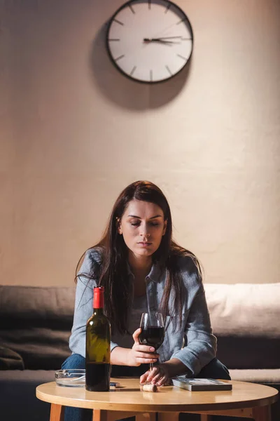 Mujer deprimida sosteniendo un vaso de vino tinto mientras está sentada sola cerca de la botella y el marco de fotos en la mesa - foto de stock