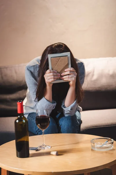 Donna depressa volto oscuro con cornice fotografica mentre seduto vicino bottiglia e bicchiere di vino rosso sul tavolo — Foto stock