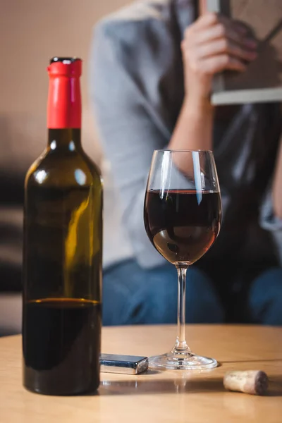 Vaso y botella de vino tinto cerca de una mujer adicta al alcohol sobre un fondo borroso - foto de stock