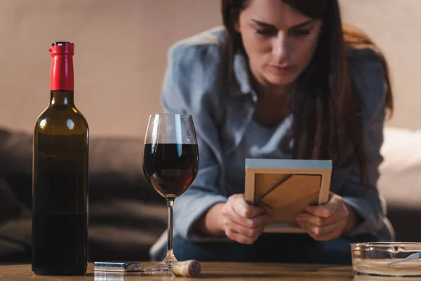 Mujer deprimida mirando el marco de la foto mientras está sentada cerca de la botella y el vaso de vino tinto, fondo borroso - foto de stock