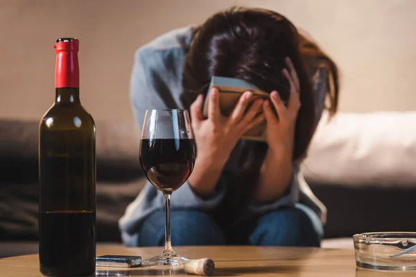 Bottle and glass of red wine near frustrated woman obscuring face with photo frame on blurred foreground — Stock Photo