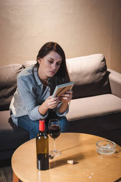 Femme bouleversée regardant cadre photo tout en étant assis à la table avec bouteille et verre de vin rouge — Photo de stock