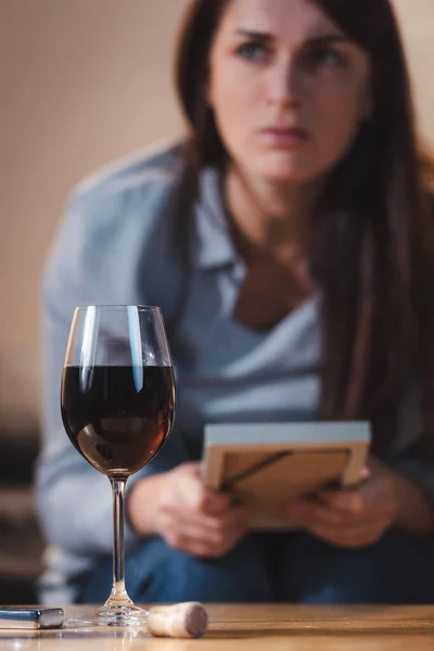 Solitário, mulher viciada em álcool segurando moldura da foto enquanto sentado perto de copo de vinho tinto, fundo borrado — Fotografia de Stock