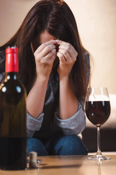 Mujer deprimida y adicta que oscurece la cara con las manos mientras sostiene el anillo de bodas cerca del vino tinto en un primer plano borroso - foto de stock