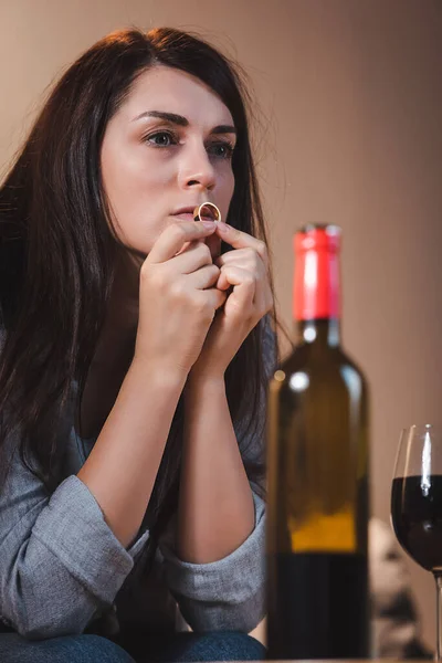 Frustrated woman holding wedding ring and looking away near bottle and glass of red wine on blurred foreground — Stock Photo