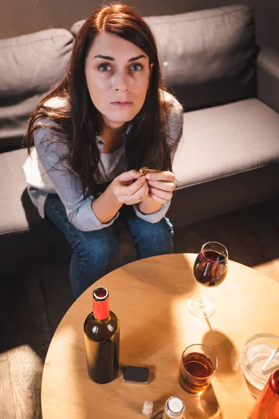 Vista ad alto angolo della donna depressa guardando la fotocamera mentre seduto vicino a bevande alcoliche e tenendo fede alla fede nuziale — Foto stock