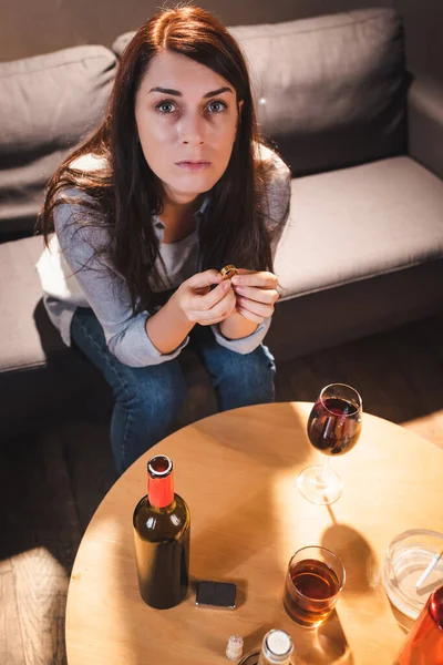 High angle view of depressed woman looking at camera while sitting near alcohol drinks and holding wedding ring — Stock Photo