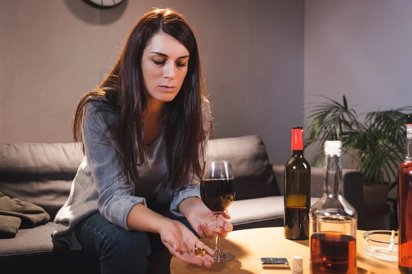 Frustrated woman holding wedding ring and glass of red wine near bottles on blurred foreground — Stock Photo