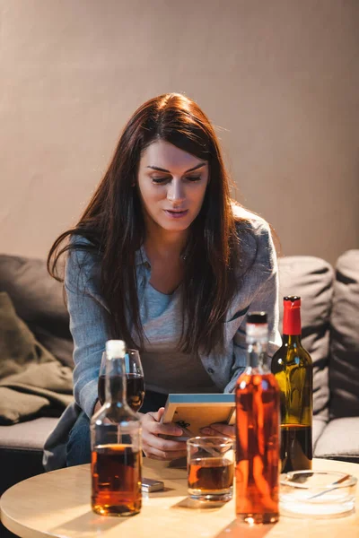 Betrunkene Frau hält Fotorahmen neben Glas und Alkoholflaschen im verschwommenen Vordergrund — Stockfoto