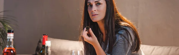 Depressed alcohol-addicted woman looking at camera while holding wedding ring near bottles on blurred foreground, banner — Stock Photo