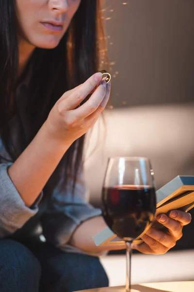 Vista ritagliata della donna che tiene cornice foto e fede nuziale vicino al bicchiere di vino rosso in primo piano sfocato — Foto stock