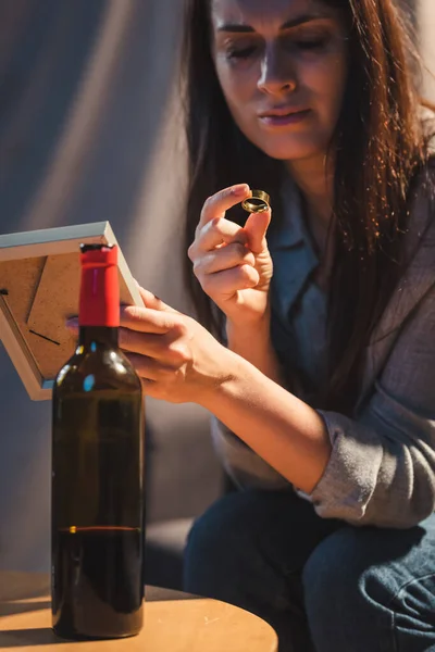 Femme déprimée tenant cadre photo et bague de mariage près de la bouteille de vin rouge — Photo de stock