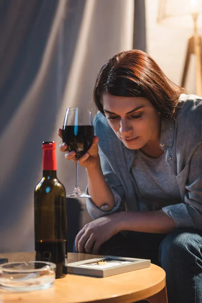 Mujer triste y solitaria mirando el marco de la foto mientras sostiene una copa de vino tinto - foto de stock