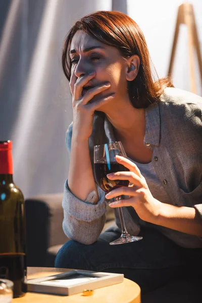 Mujer borracha llorando mientras sostiene un vaso de vino tinto cerca del marco de la foto y el anillo de boda en la mesa - foto de stock