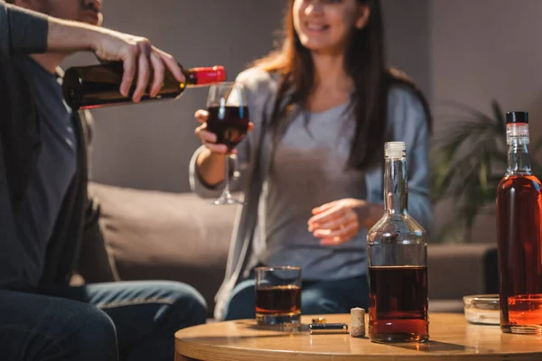 Vista ritagliata del marito versando vino nel bicchiere di moglie vicino a bottiglie di alcol sul tavolo, sfondo sfocato — Foto stock