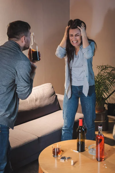 Depressed, drunk woman touching head while standing with closed eyes near husband with bottle of whiskey — Stock Photo