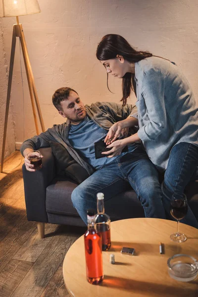 Frau zeigt betrunkenem Mann, der mit Whiskey auf Sofa sitzt, Münzen und leere Geldbörse — Stockfoto