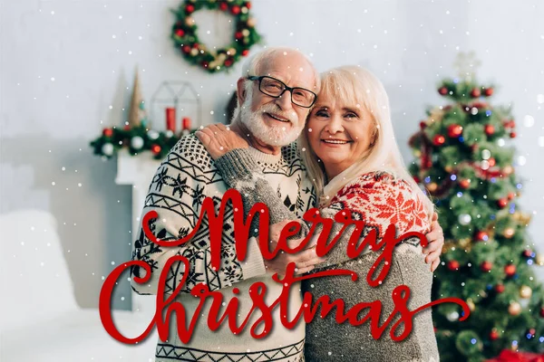 Happy senior couple embracing while looking at camera near merry christmas lettering and decorations on blurred background — Stock Photo