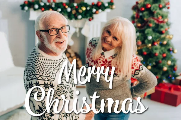 Joyeux homme et femme âgés regardant caméra près joyeux Noël lettrage et décorations sur fond flou — Photo de stock
