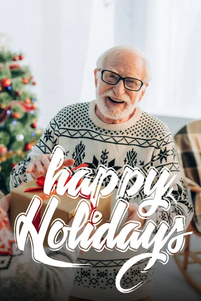 Fröhlicher Senior erhält Weihnachtsgeschenk von Frau in der Nähe frohe Feiertage Schriftzug zu Hause — Stockfoto