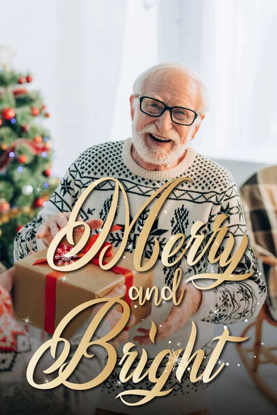 Homem sênior feliz recebendo presente de Natal da esposa perto de letras alegres e brilhantes em casa — Fotografia de Stock
