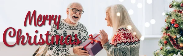 Hombre mayor emocionado presentando regalo a esposa feliz cerca de tipografía feliz de Navidad, bandera - foto de stock