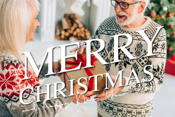 Aufgeregter Senior mit Brille nimmt Geschenk von lächelnder Frau nahe froher Weihnachten und frohem neuen Jahr entgegen — Stockfoto