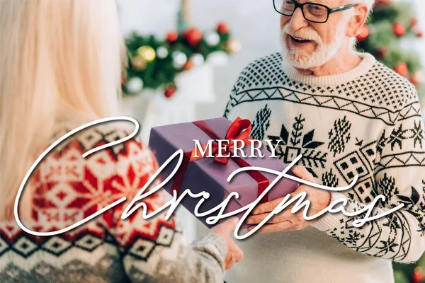 Senior überreicht Frau Geschenkschachtel in der Nähe von frohem Weihnachten Schriftzug auf unscharfem Vordergrund — Stockfoto