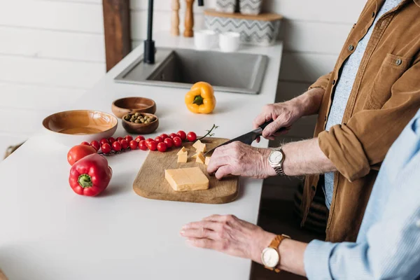 Cropped view of elderly wife near husband cutting cheese on chopping board in kitchen on blurred background — Stock Photo