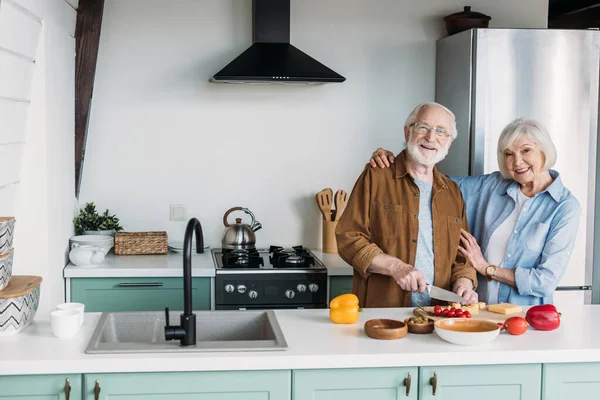 Усміхнена старша дружина дивиться на камеру, обіймаючи чоловіка готуючи вечерю на кухні — стокове фото