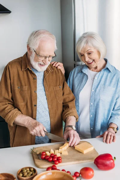 Feliz anciana esposa cerca de marido corte queso en la tabla de cortar cerca de verduras en la cocina - foto de stock