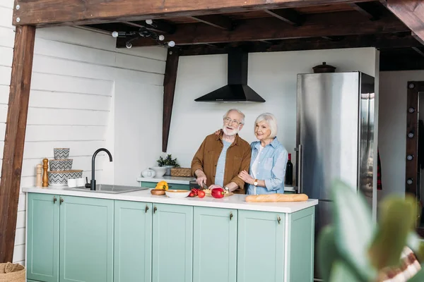 Sonriente mujer mayor abrazando al hombre cortando queso en la mesa con verduras y baguette en la cocina en primer plano borroso - foto de stock